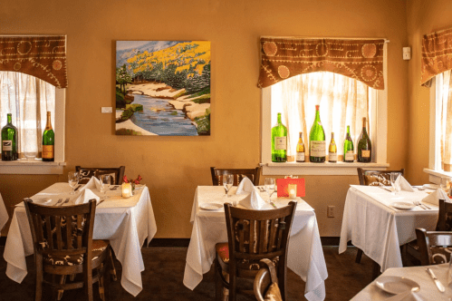 Cozy restaurant interior with white tablecloths, wine bottles on shelves, and a landscape painting on the wall.